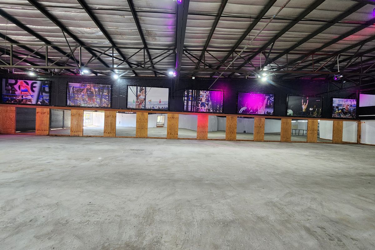 Spacious industrial-style gym area featuring exposed steel roof trusses, concrete flooring, and a long wall of mirrors, suitable for fitness or studio use.
