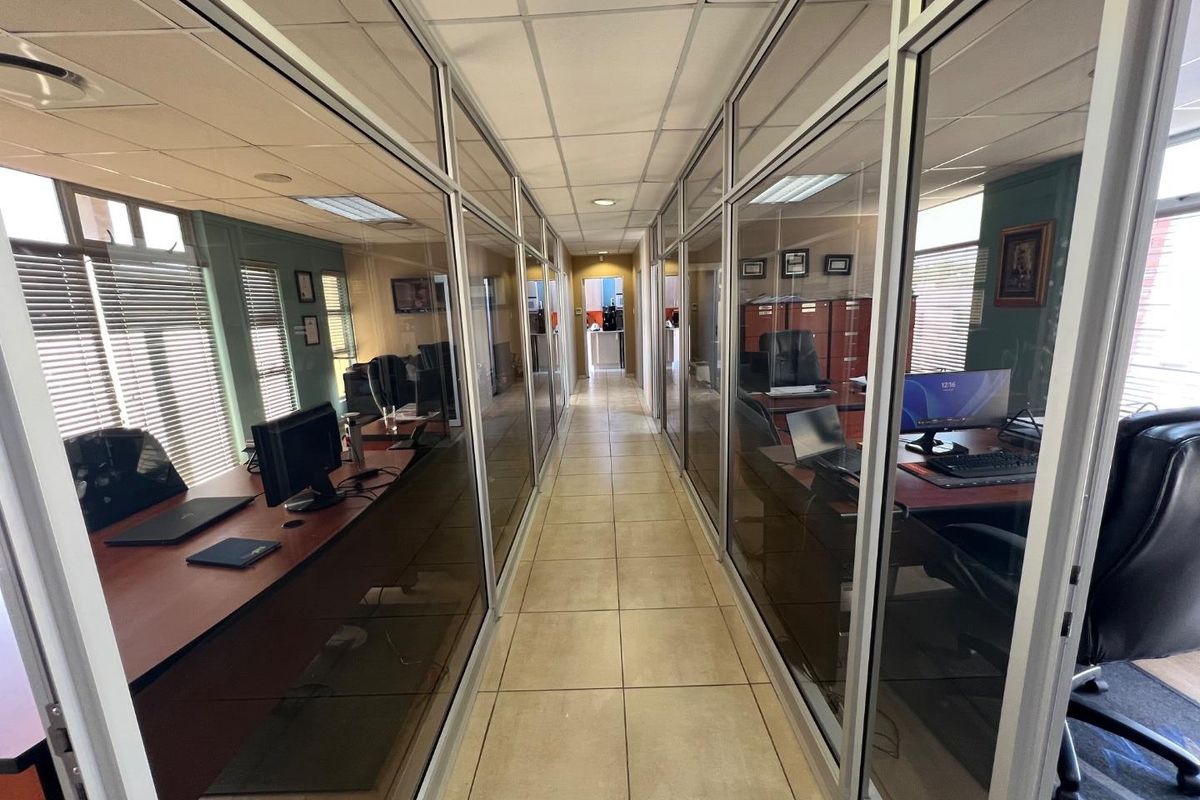 A commercial office hallway featuring glass-partitioned office suites with tiled flooring and suspended ceiling panels.