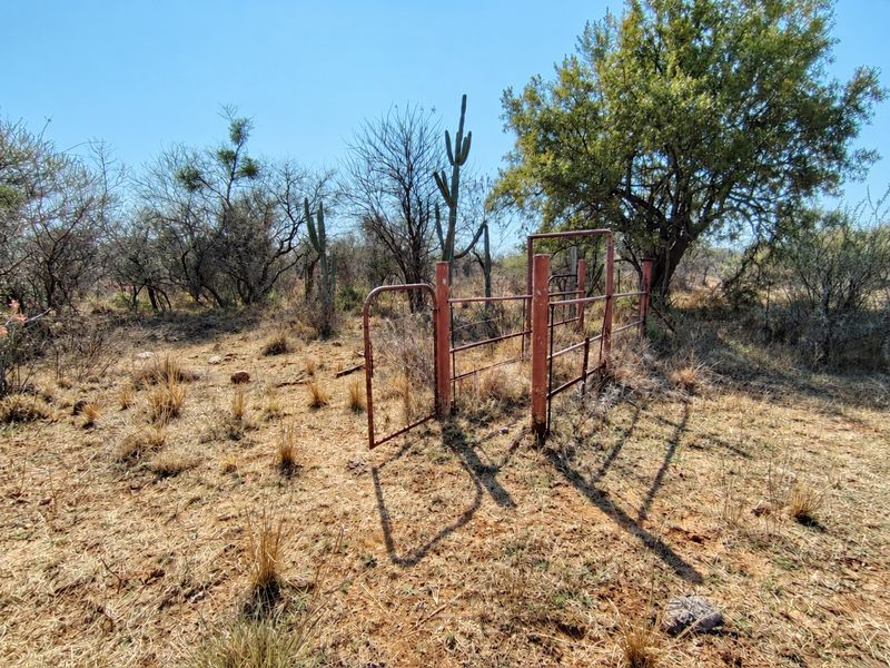 A view of a vacant commercial land plot featuring a weathered metal gate and wire fencing amidst natural desert vegetation.