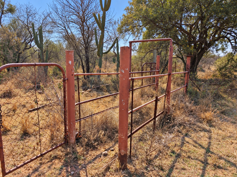A view of a vacant land plot featuring weathered metal fencing and gate posts set within a natural, dry landscape with native vegetation.