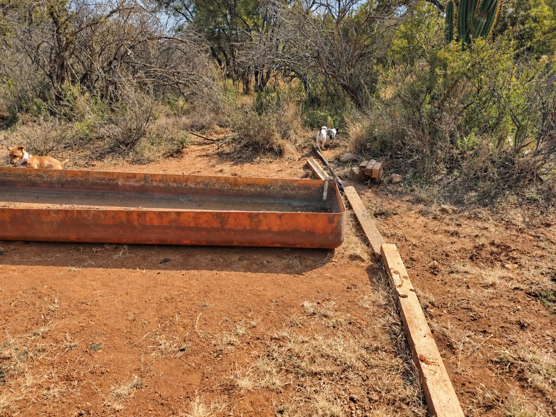 An outdoor area featuring a weathered metal water trough and basic irrigation infrastructure set within a natural, undeveloped landscape.