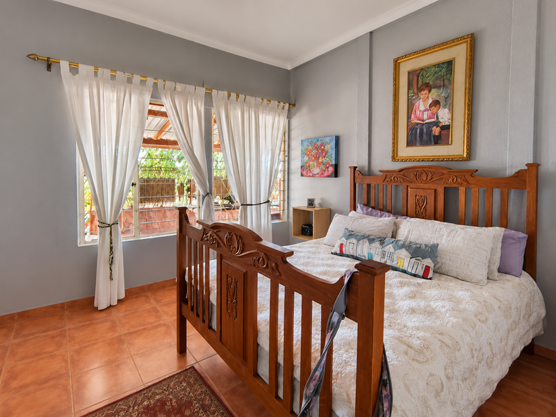 A well-maintained bedroom featuring terracotta floor tiles, neutral grey walls, and a window with security bars, offering a comfortable and functional living space.