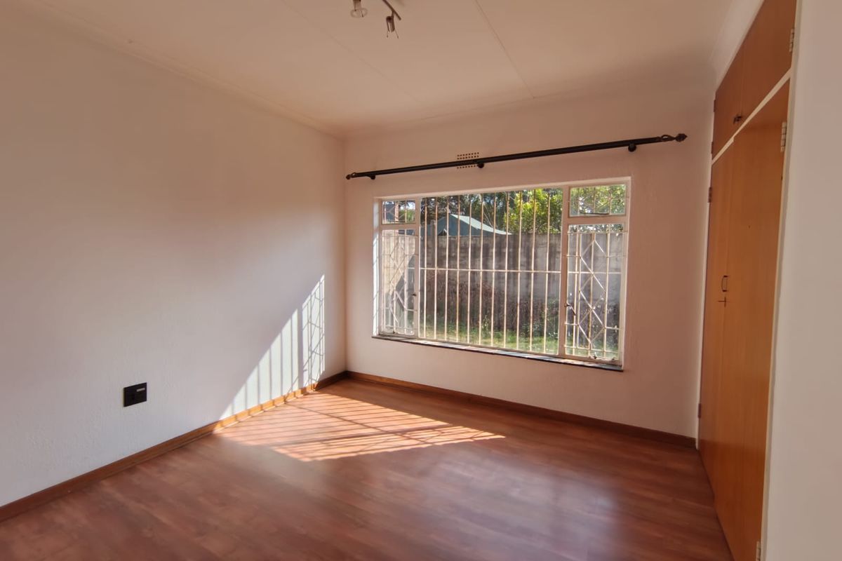 A bright, empty bedroom featuring laminate flooring, a built-in wooden wardrobe, and a large window fitted with security bars.