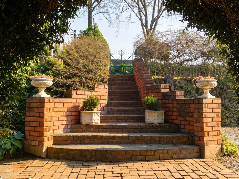 A charming garden setting featuring a brick staircase with retaining walls, leading through a metal gate, framed by lush greenery and paved pathways.