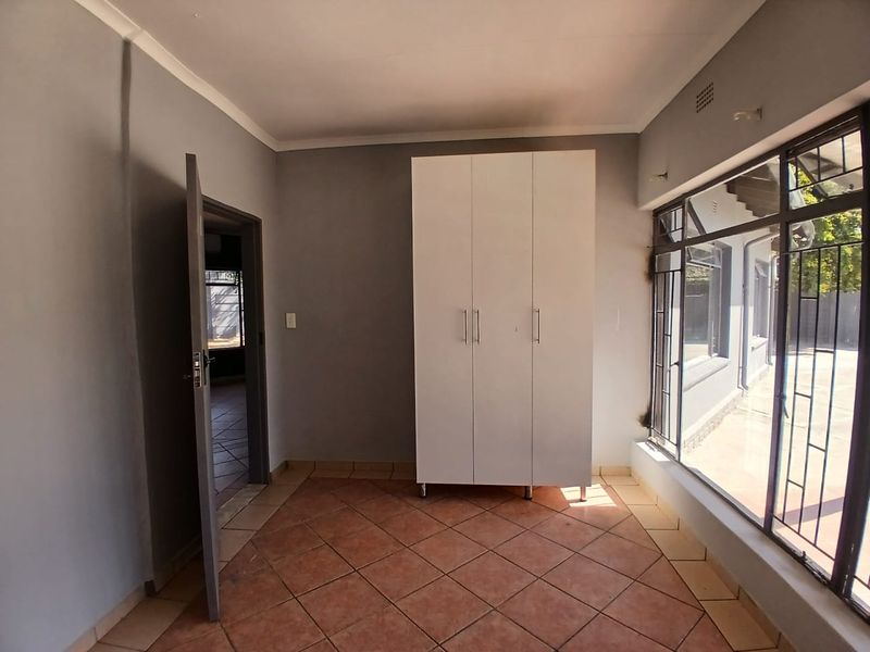 A functional office space featuring tiled flooring, a built-in cupboard, and large windows with security bars, offering ample natural light.