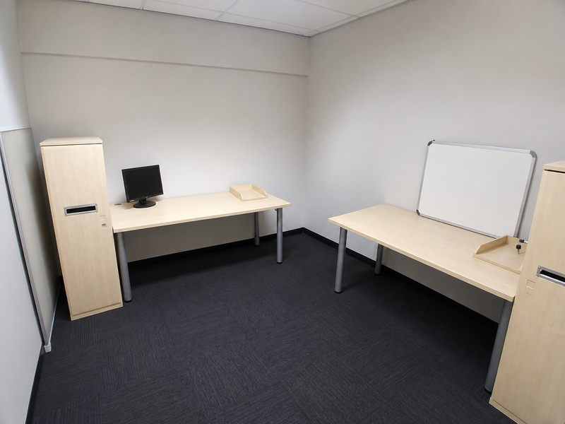 A clean, functional office space featuring neutral walls, dark carpet tiles, a suspended ceiling, and integrated storage cabinetry.
