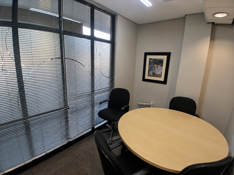 A professional office space featuring large floor-to-ceiling windows with venetian blinds, recessed ceiling lighting, and neutral carpeted flooring.