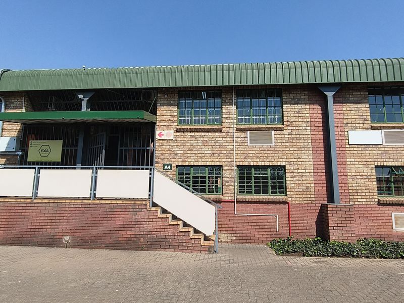 Front exterior view of an industrial brick building featuring a metal roof, security-barred windows, and a paved entrance area with stairs.