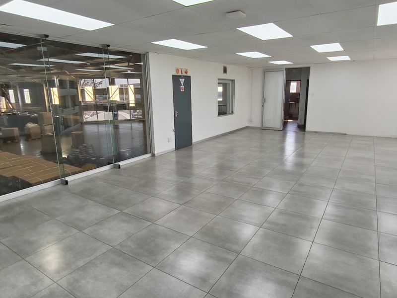 Spacious, open-plan industrial office area featuring modern tiled flooring, suspended ceilings with recessed lighting, and a large glass partition wall overlooking the warehouse space.
