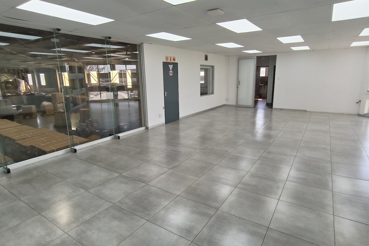 Spacious, open-plan industrial office area featuring modern tiled flooring, suspended ceilings with recessed lighting, and a large glass partition wall overlooking the warehouse space.