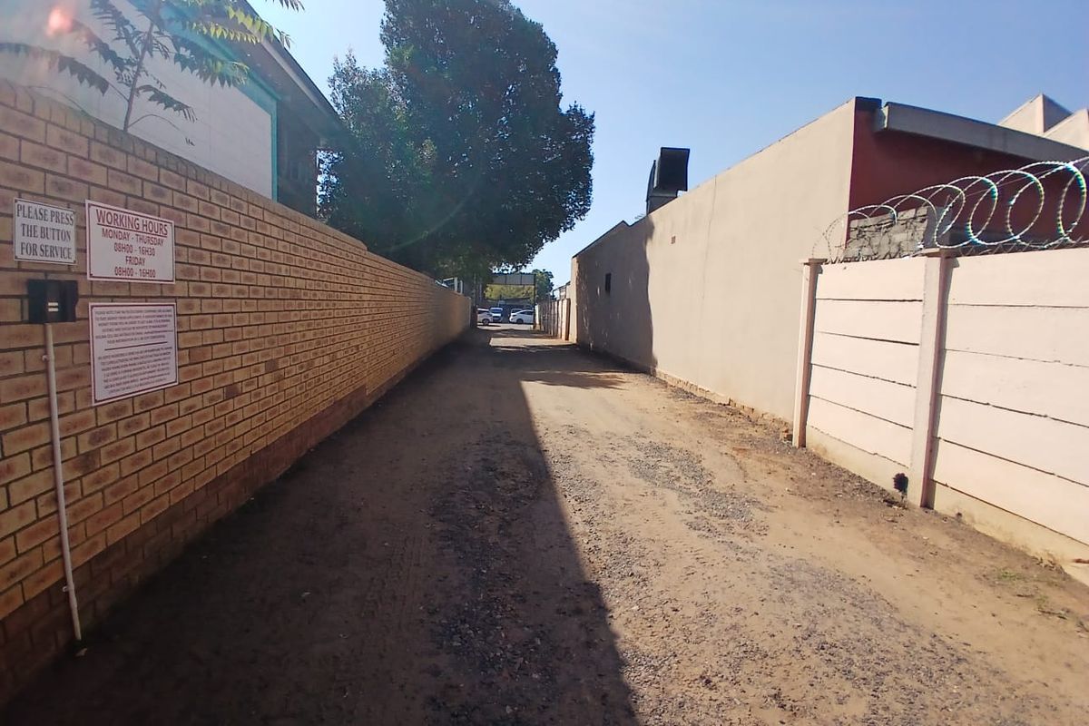 A narrow, unpaved rear access driveway enclosed by brick and precast concrete boundary walls with security razor wire.