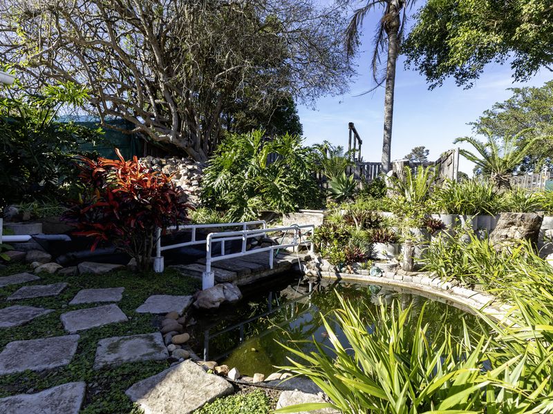 A landscaped garden area featuring a stone pathway, a small pond with a pedestrian bridge, and established greenery with retaining wall structures.