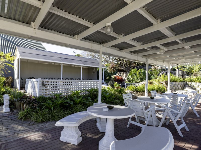 A covered outdoor patio area featuring brick paving, a corrugated metal roof structure, and a landscaped garden setting.
