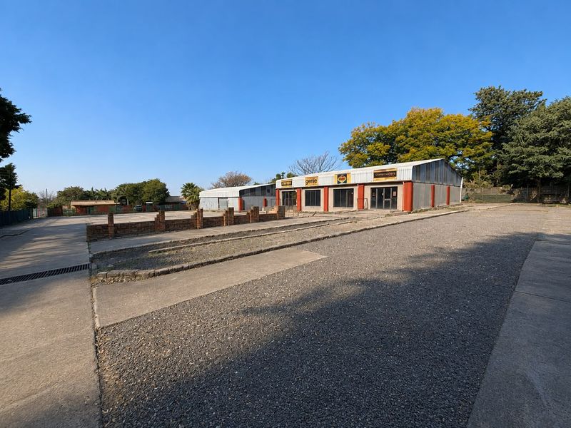 Front view of an industrial property featuring a large concrete parking area, brick boundary wall, and metal-clad warehouse buildings with multiple access points.