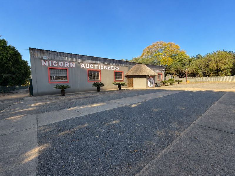 Front exterior view of an industrial property featuring a corrugated metal facade, thatched entrance canopy, and a large paved parking area.