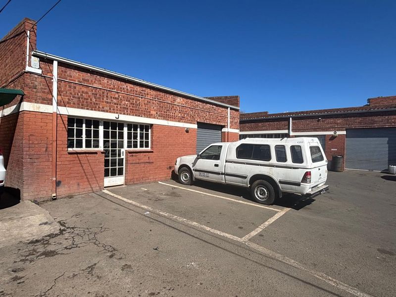 Industrial unit exterior featuring red brick facade, roller shutter doors, and dedicated asphalt parking space in a functional commercial setting.