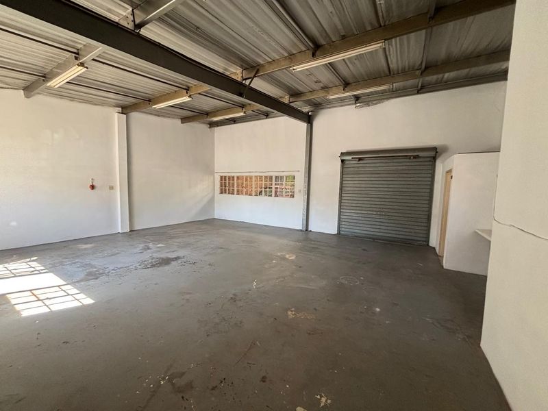 Spacious industrial utility area featuring exposed steel beams, concrete flooring, a roller shutter door, and natural light from a glass block window.