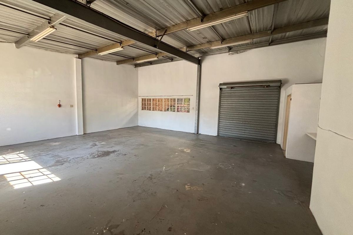 Spacious industrial utility area featuring exposed steel beams, concrete flooring, a roller shutter door, and natural light from a glass block window.