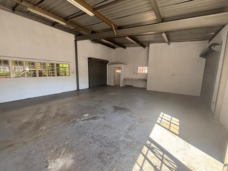 Spacious industrial utility area featuring exposed steel beams, concrete flooring, roller shutter doors, and functional fluorescent lighting, suitable for workshop or storage use.