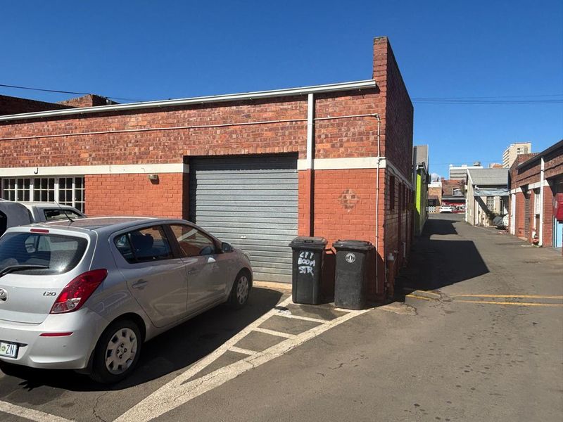 Rear exterior view of an industrial property featuring a roller shutter door and exposed brick facade with adjacent asphalt access lane.