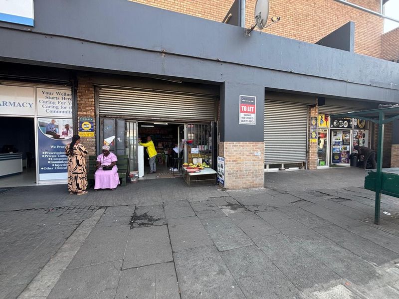 Front exterior view of a retail shop unit featuring roller shutter doors and brick facade, located along a paved sidewalk.