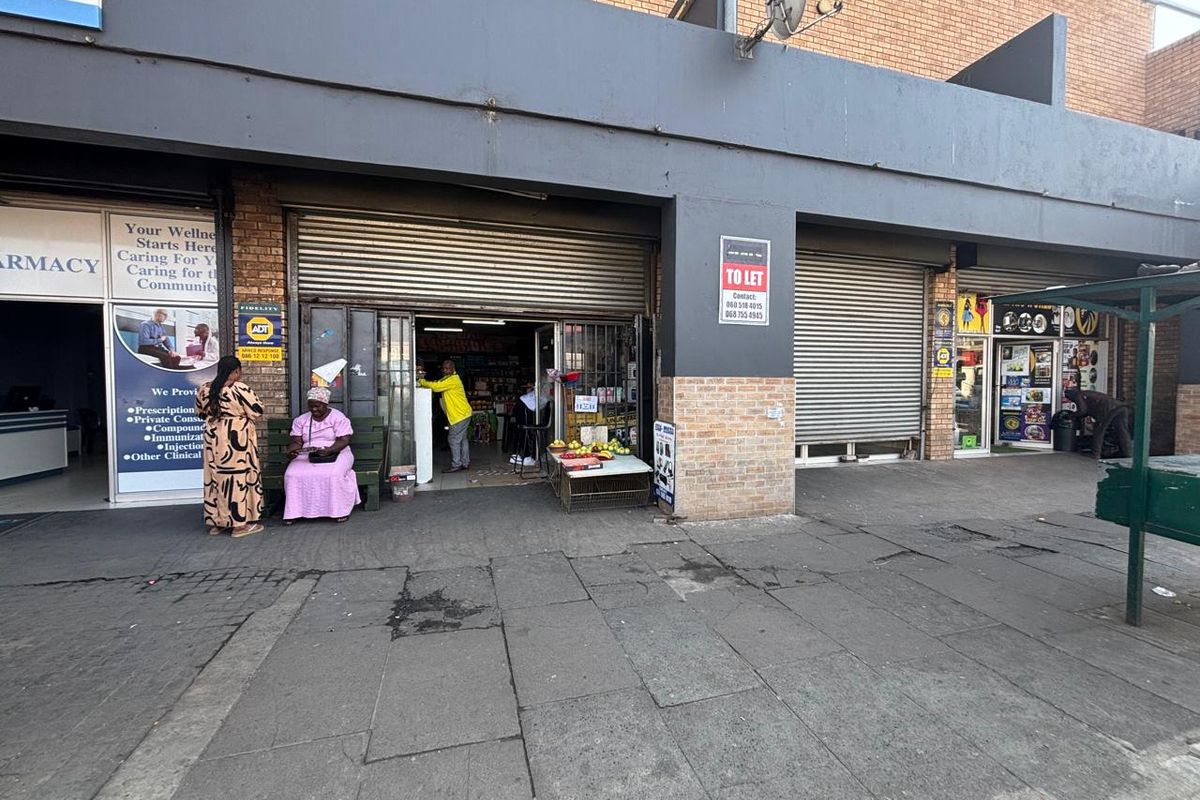 Front exterior view of a retail shop unit featuring roller shutter doors and brick facade, located along a paved sidewalk.