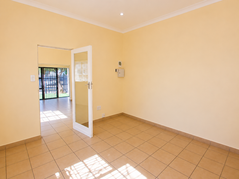A bright, empty office space featuring neutral walls, ceramic tiled flooring, and a glass-panelled door leading to an adjacent area with security gate access.