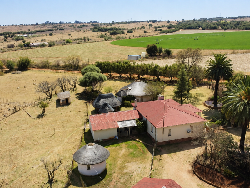 Aerial view of a farm property featuring a main residence with a corrugated metal roof, several outbuildings with thatch roofs, and surrounding agricultural land.