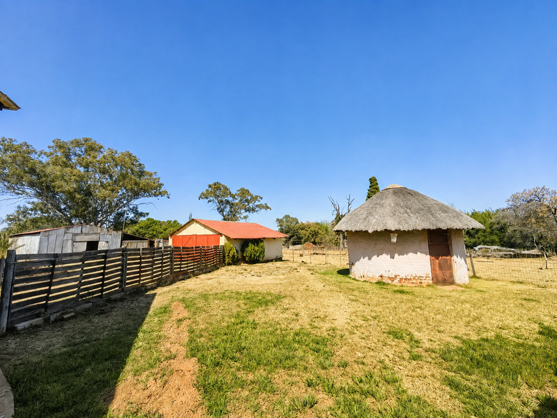 A rural farm yard featuring a traditional thatched rondavel, a corrugated iron shed, and perimeter fencing set within a dry, open landscape.