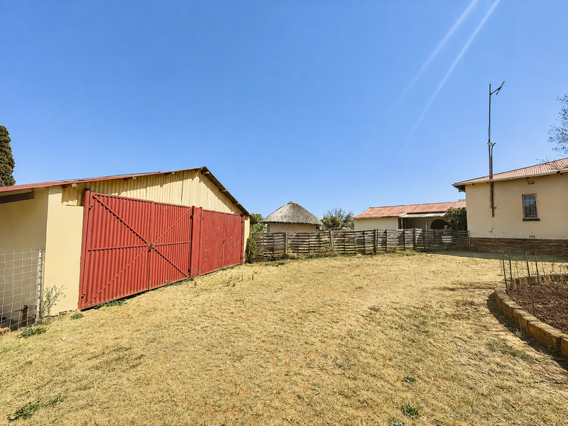 A spacious farm yard featuring a large corrugated metal barn, a traditional thatched rondavel, and established timber fencing under a clear blue sky.