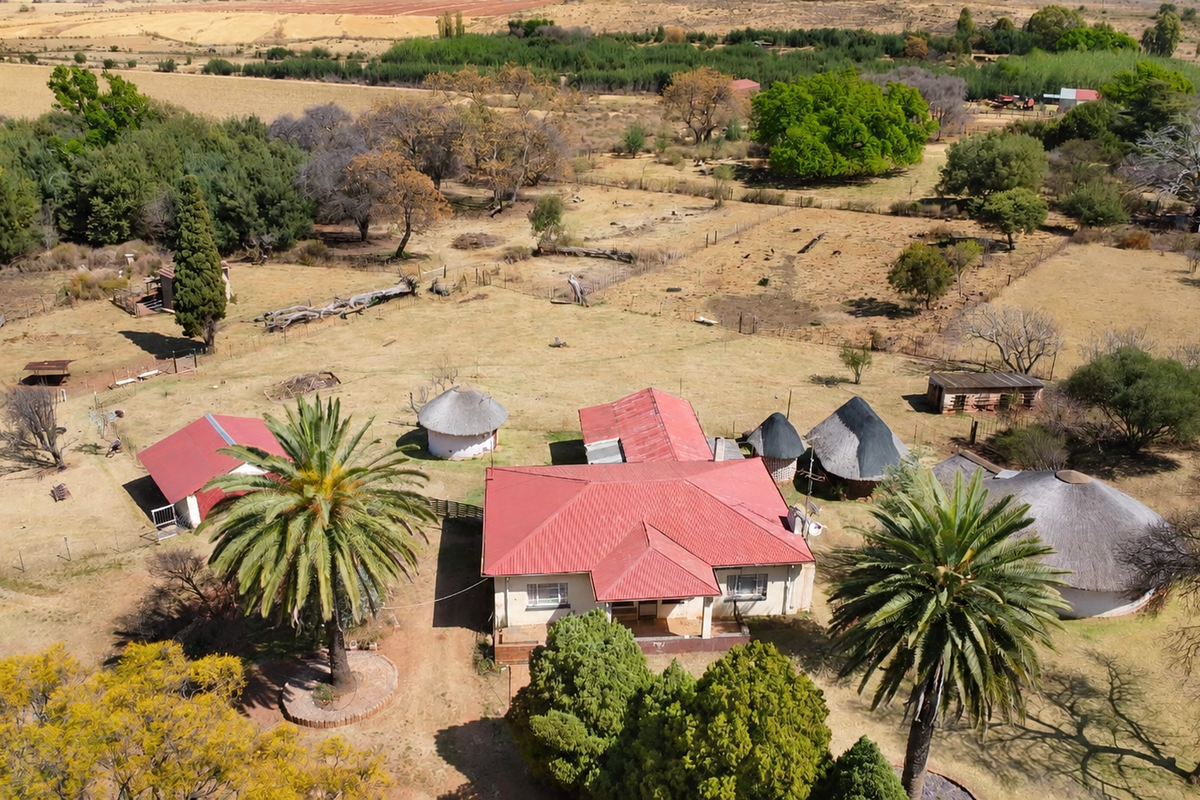 Aerial view of a farm property featuring a main residence with a metal roof, several outbuildings with thatched roofs, and surrounding agricultural land.
