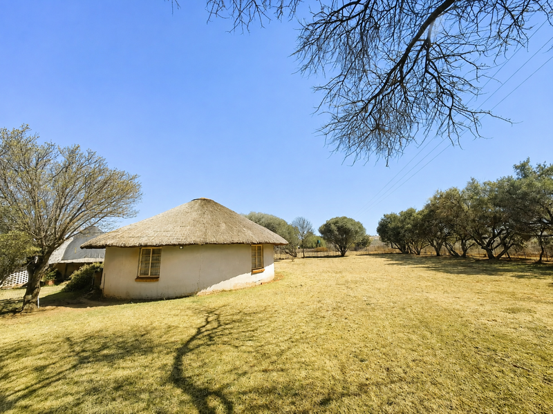 A traditional round thatched-roof cottage situated on a spacious, open farm landscape with mature trees and dry grass.