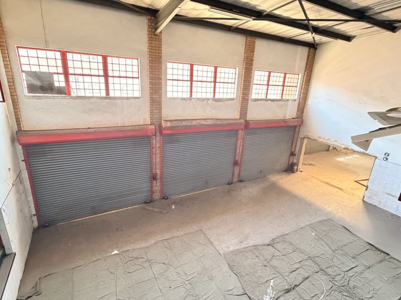 Spacious industrial utility area featuring high ceilings with exposed steel trusses, multiple roller shutter doors, and large windows providing ample natural light.