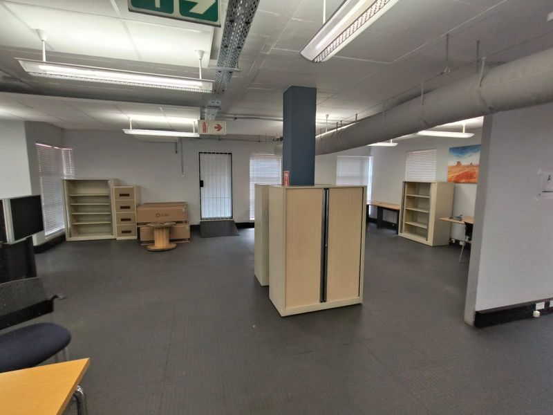 Open-plan office space featuring industrial-style exposed ceiling ducting, vinyl flooring, and natural light through multiple windows with security gates.