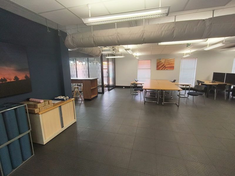 Open-plan office space featuring industrial-style exposed ductwork, suspended ceiling tiles, durable rubber flooring, and large windows with venetian blinds.