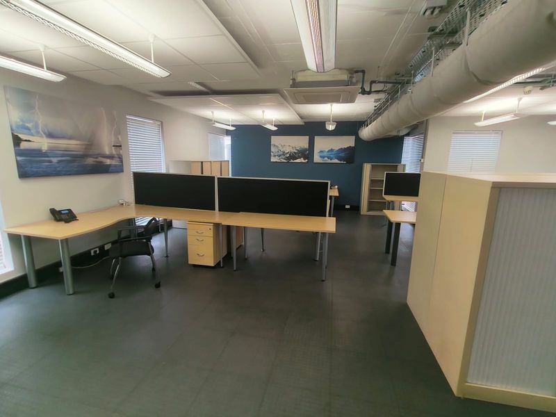 A functional open-plan office space featuring suspended ceilings, exposed industrial ducting, and large windows with venetian blinds.