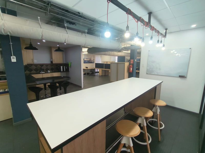 Modern open-plan office space featuring a kitchenette, exposed industrial ceiling ducting, pendant lighting, and a wall-mounted whiteboard for collaborative work.
