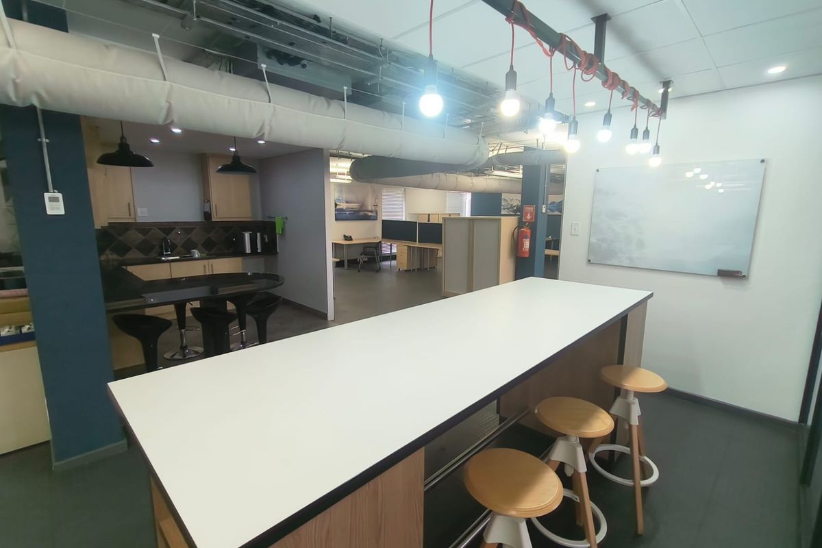 Modern open-plan office space featuring a kitchenette, exposed industrial ceiling ducting, pendant lighting, and a wall-mounted whiteboard for collaborative work.
