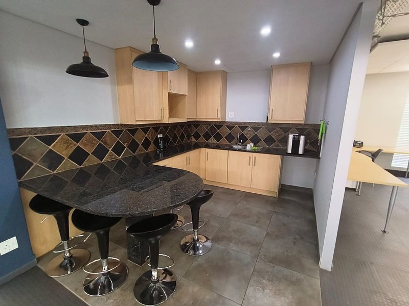 A functional office kitchenette featuring light wood cabinetry, dark granite countertops, a decorative tiled backsplash, and integrated sink with modern pendant lighting.