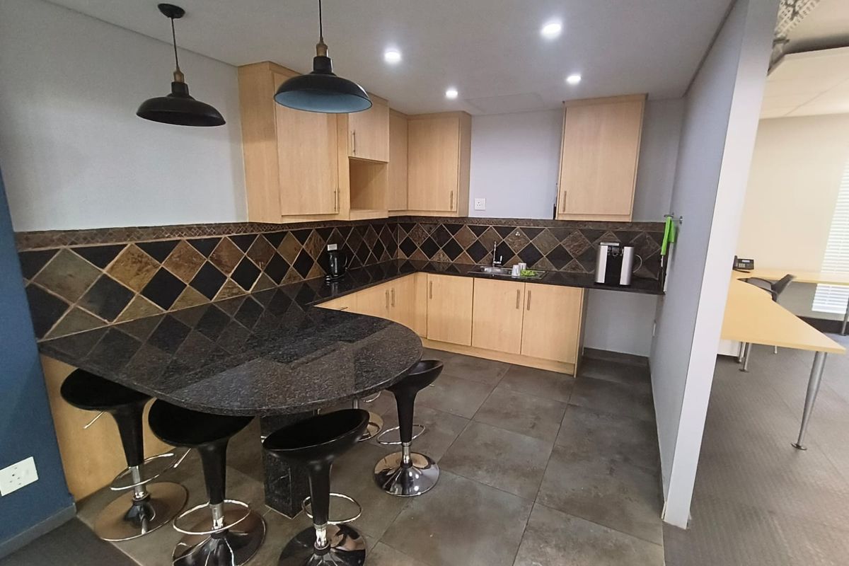 A functional office kitchenette featuring light wood cabinetry, dark granite countertops, a decorative tiled backsplash, and integrated sink with modern pendant lighting.