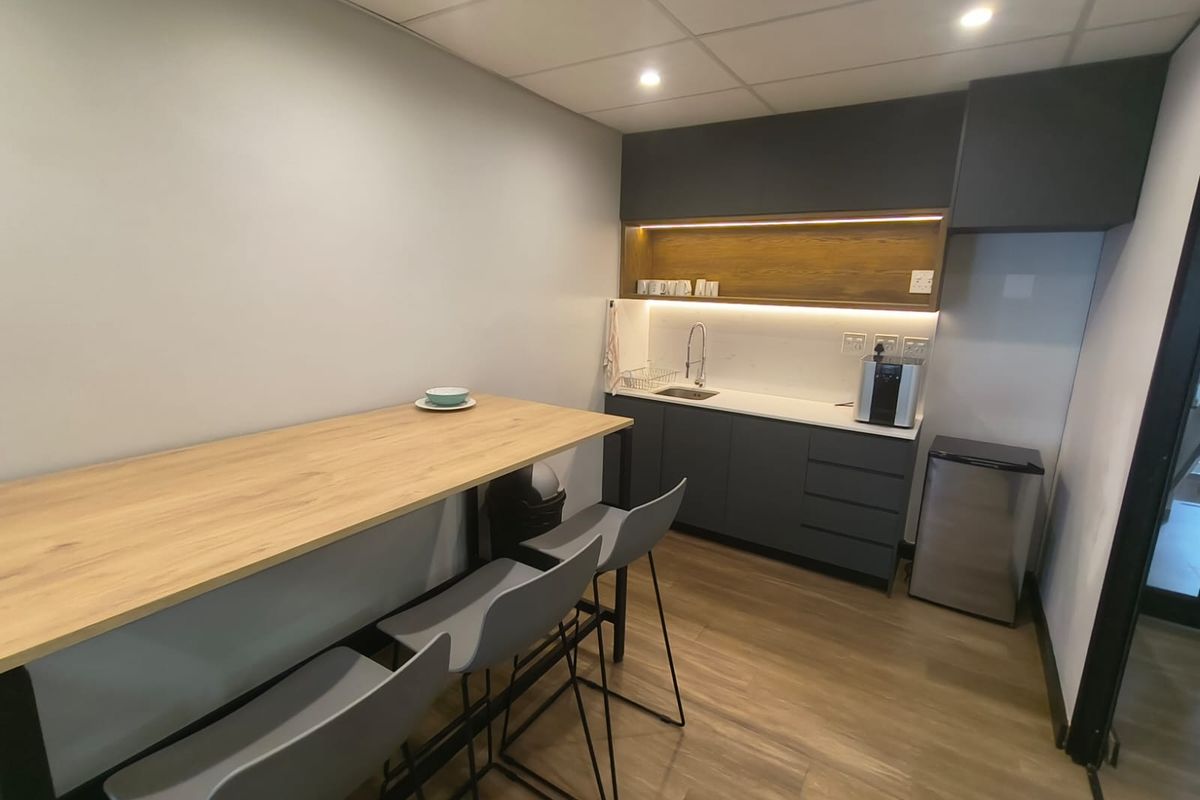 A modern office kitchenette featuring dark grey cabinetry, a light wood-look countertop, integrated sink, and recessed ceiling lighting.