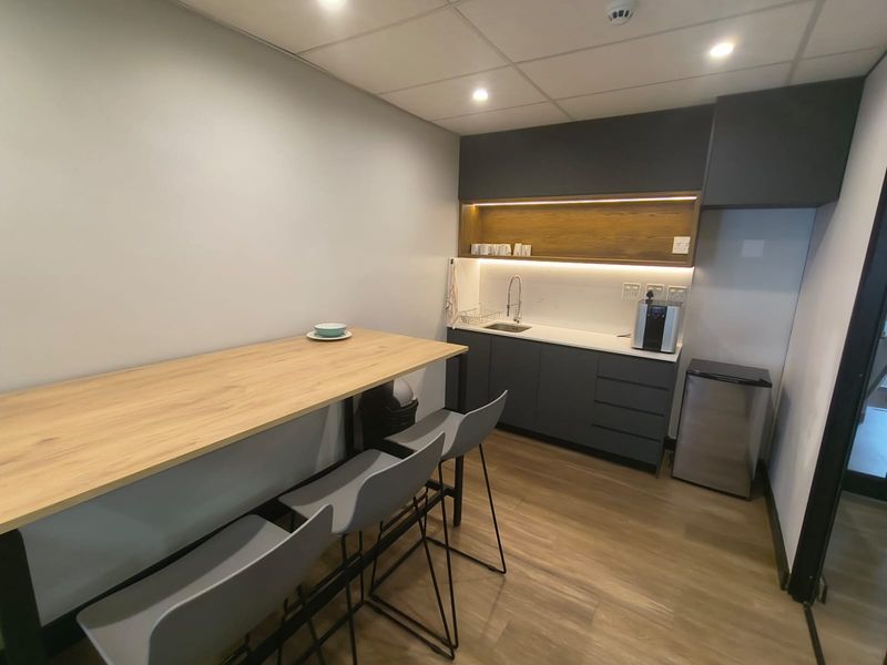A modern office kitchenette featuring dark grey cabinetry, a light wood-look countertop, integrated sink, and recessed ceiling lighting.