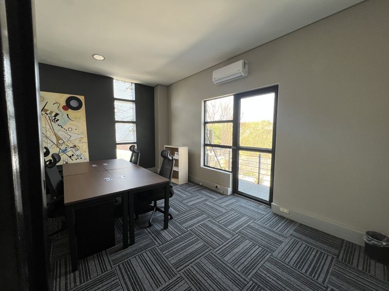 A modern office space featuring carpet tiles, recessed lighting, a wall-mounted air conditioning unit, and access to an external balcony through a glass door.