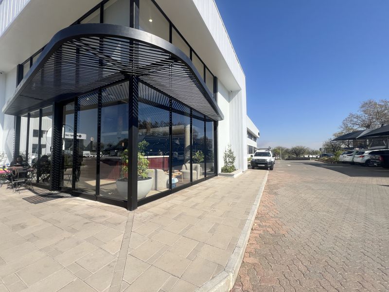 Modern office building exterior featuring a prominent glass corner facade, sleek black metal architectural detailing, and paved pedestrian and vehicle access areas.