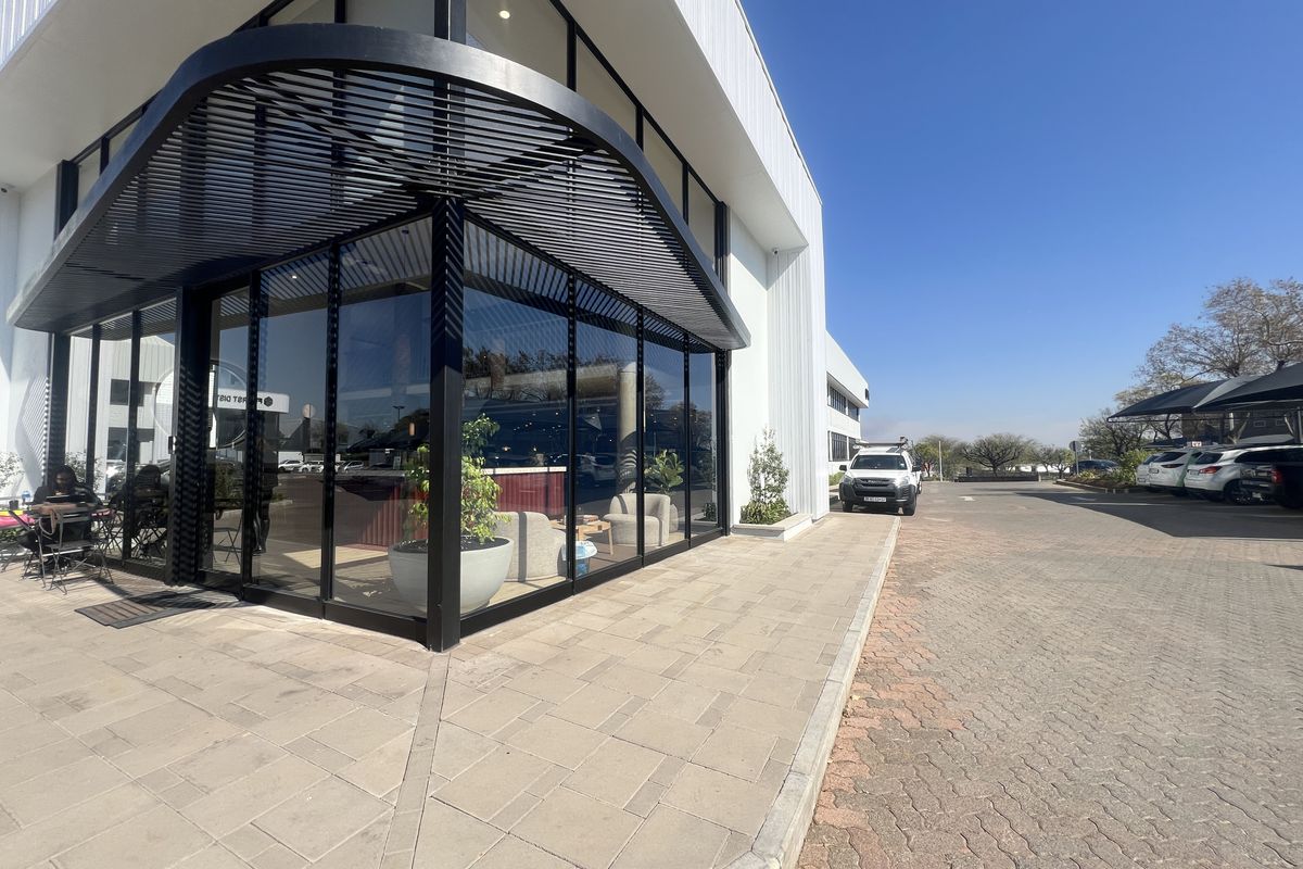 Modern office building exterior featuring a prominent glass corner facade, sleek black metal architectural detailing, and paved pedestrian and vehicle access areas.