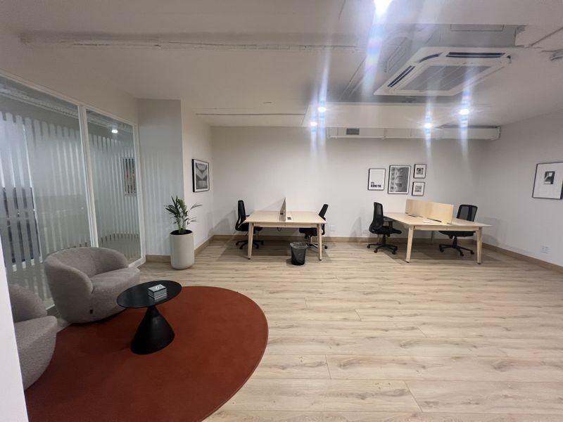 A modern, open-plan office space featuring light wood-look flooring, a glass partition wall, and a recessed ceiling air conditioning unit.