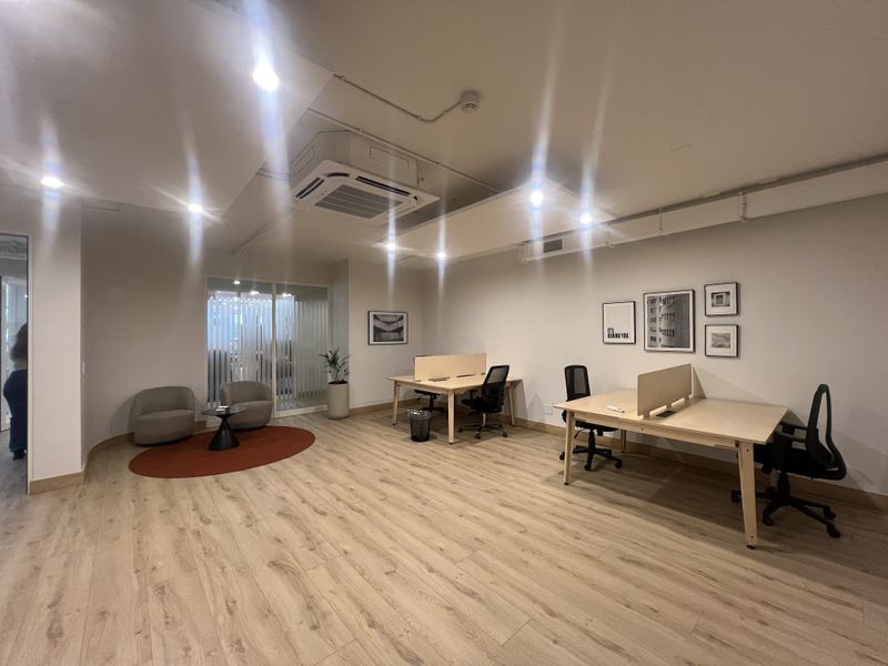 Modern open-plan office space featuring light wood-look flooring, recessed ceiling lighting, and a central ducted air conditioning unit with glass partition access.