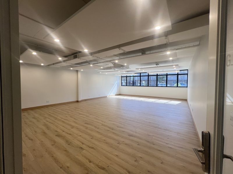 A bright, open-plan office space featuring modern laminate wood flooring, suspended acoustic ceiling panels with recessed lighting, and large windows providing natural light.