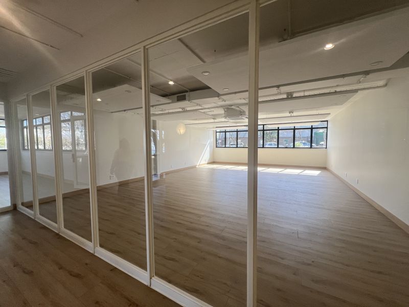 A bright, modern, and empty office space featuring glass partitions, laminate flooring, and large windows providing ample natural light.