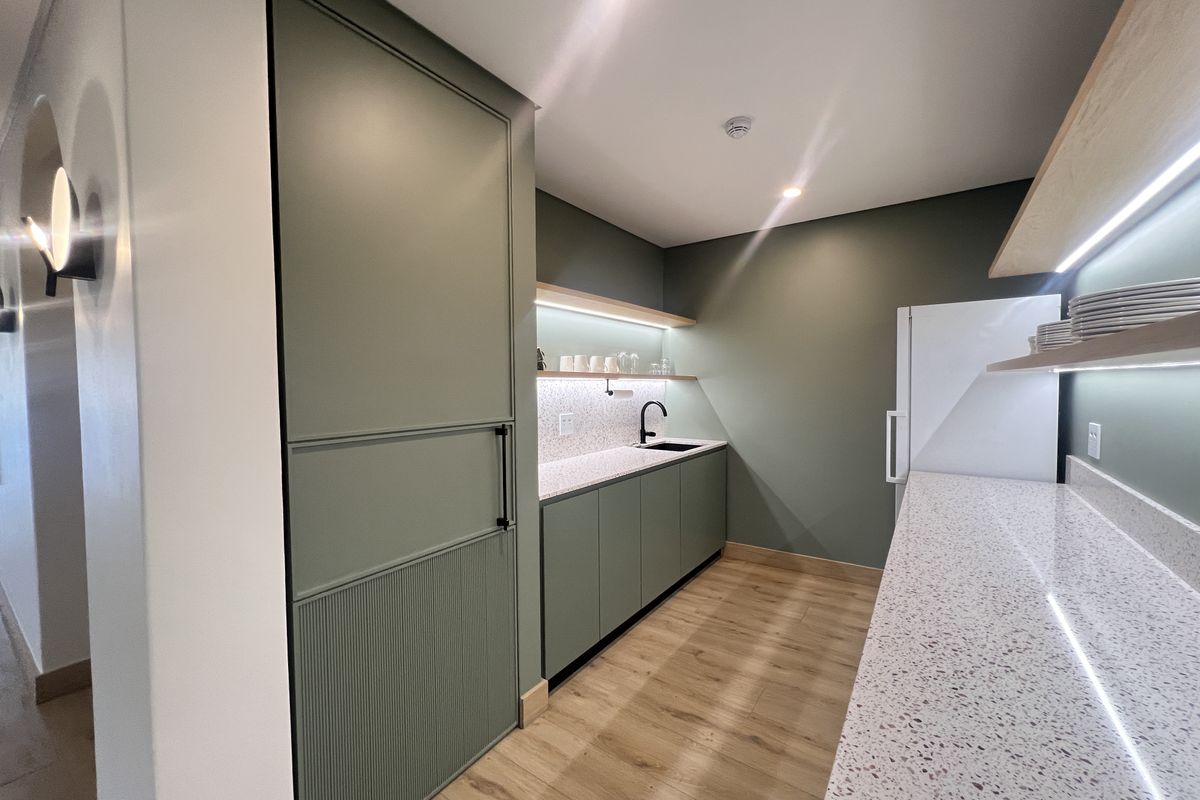 Modern office kitchenette featuring sage green cabinetry, terrazzo countertops, floating shelves with integrated LED lighting, and high-quality wood-look flooring.